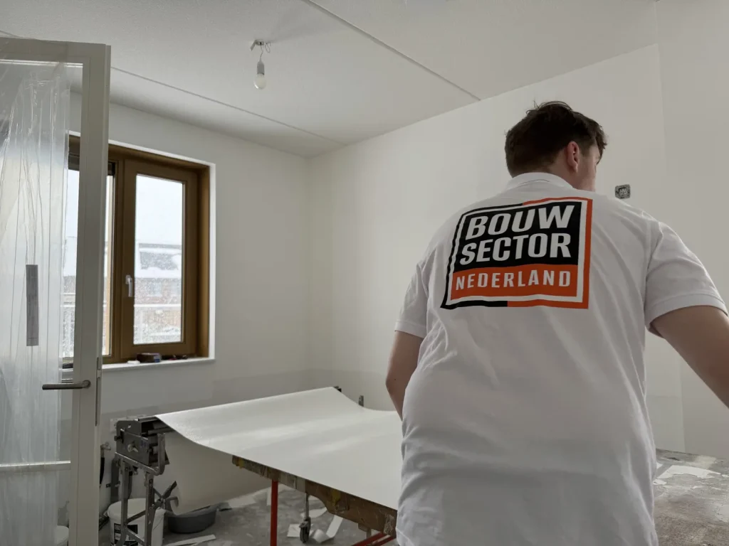 Behanger in een wit shirt met het Bouwsector-logo staat bij de plaktafel met een baan behang in een lege kamer