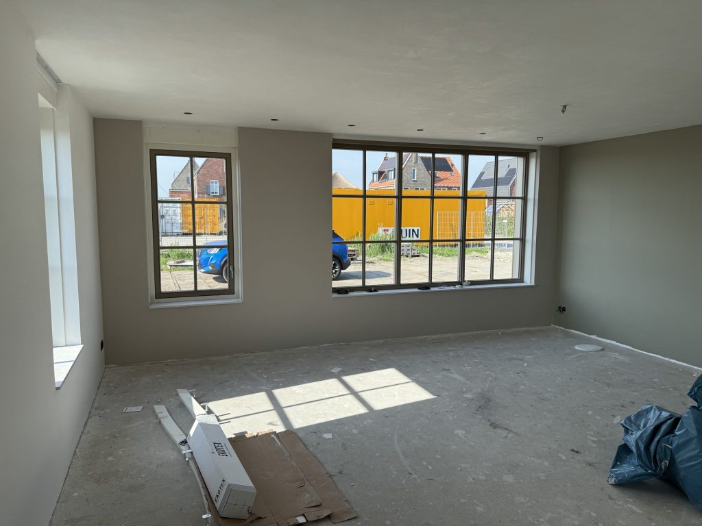 Woonkamer van een nieuw huis met grijs gesausde wanden, grote sprossenramen en zonlicht op de kale vloer