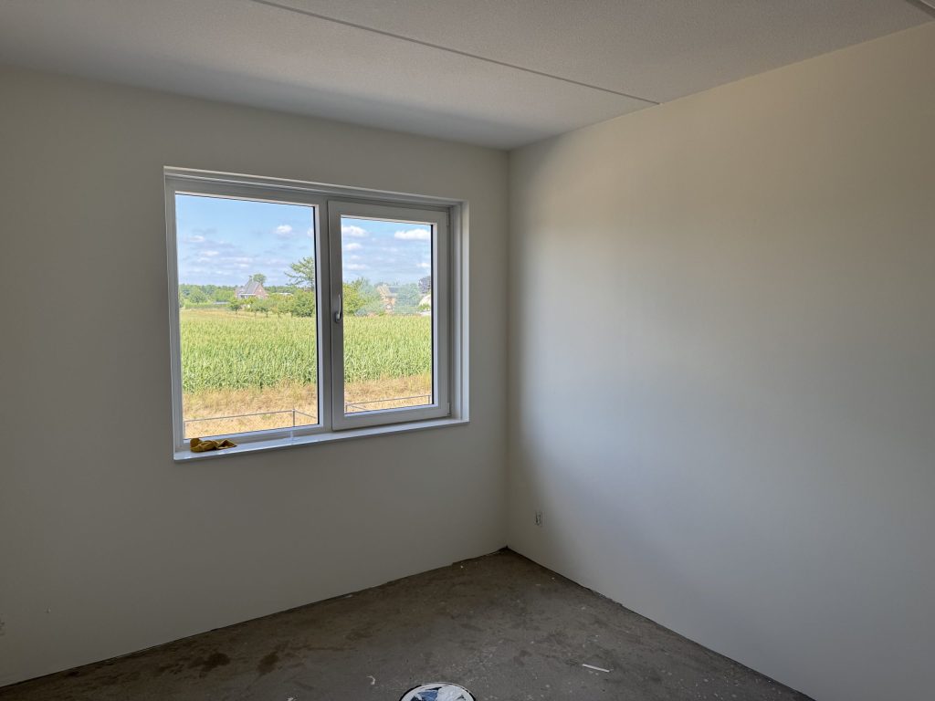 Lege slaapkamer in een nieuwbouwhuis met gladde lichte wanden en een raam dat uitkijkt over een maisveld