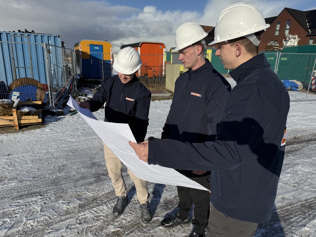 Drie vakmensen van Bouwsector Nederland met witte helm bekijken samen een bouwtekening op een besneeuwde bouwplaats